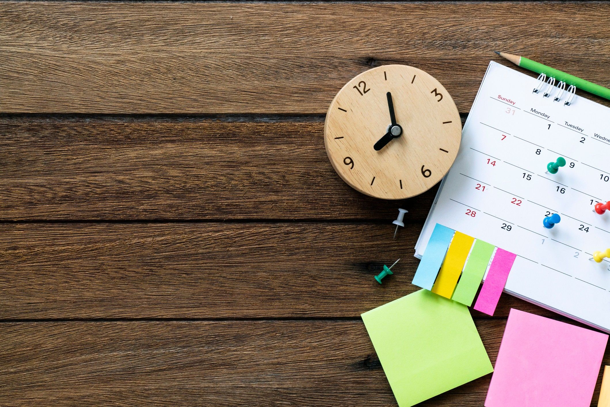 A wooden table with a calendar, pencil, post-it notes and wooden clock.
