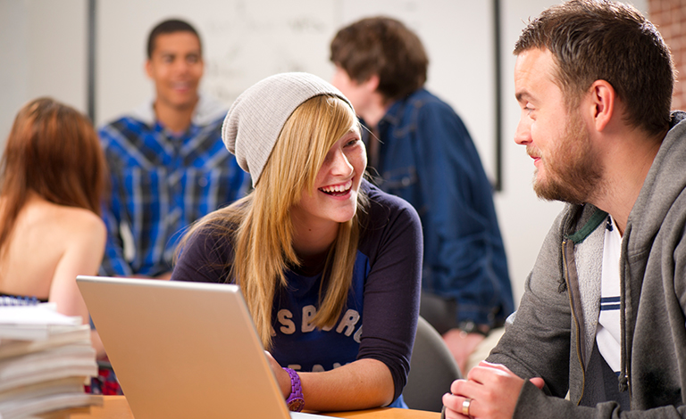 A young woman sits at a laptop laughing with a man.