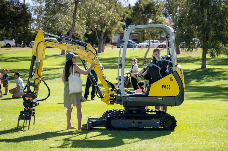 A boy on a yellow digger with his mum looking on
