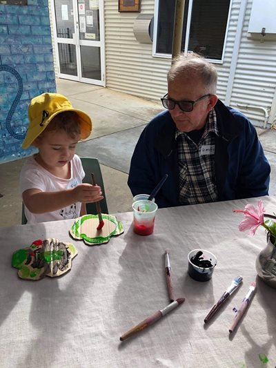 A young child paints a cardboard flower next to an elderly man. 