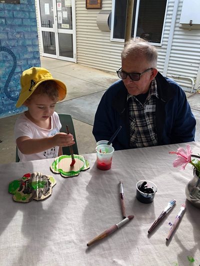 A young child paints a cardboard flower next to an elderly man.
